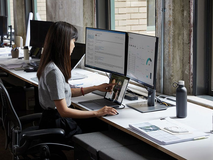 zu sehen ist eine dunkelhaarige Frau an einem Schreibtisch vor zwei Monitoren. auf denen Office Anwendungen gezeigt werden. Thema ist die Datenkompetenz im Zuge der Digitalisierung. Bild: Microsoft