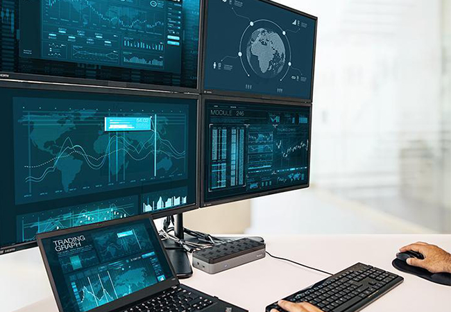 Zu sehen ist ein Arbeitsplatz mit Laptop, vier Monitoren und einer Dockingstation für Laptops, Hände bedienen Maus und Tastatur. Small Data Bild: Targus