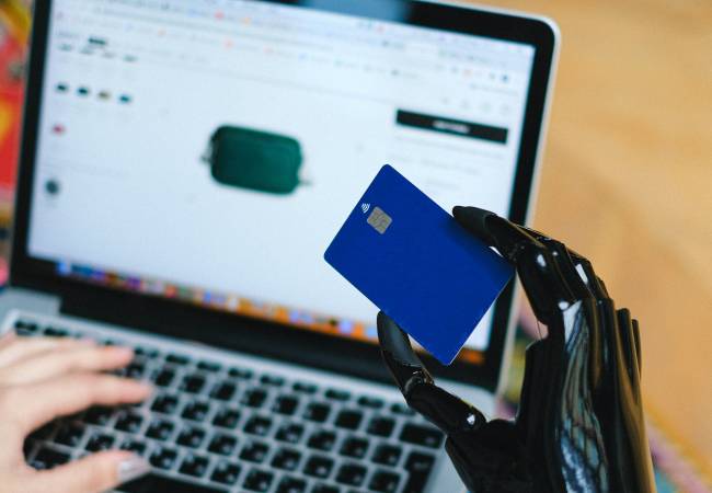 Zu sehen ist eine Roboterhand, die eine Kreditkarte hält, dahinter ein Laptop - es geht um KI-Angriffe. Bild: Pexels/Anna Shvets