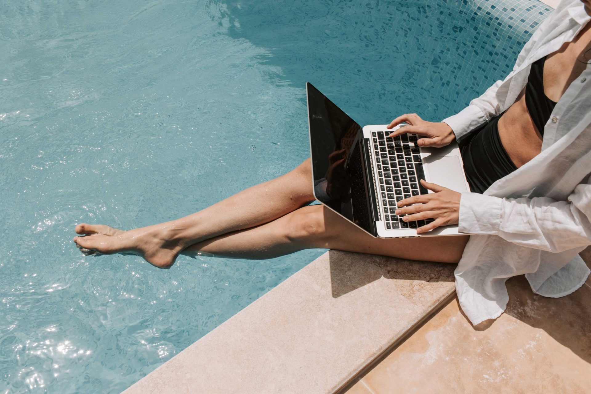 zu sehen ist der Körper einer Frau. Sie sitzt mit ihrem Laptop am Pool und hat die Füße im Wasser. Thema ist das Outdoor-Homeoffice. Bild: Pexels/Vlada Karpovich